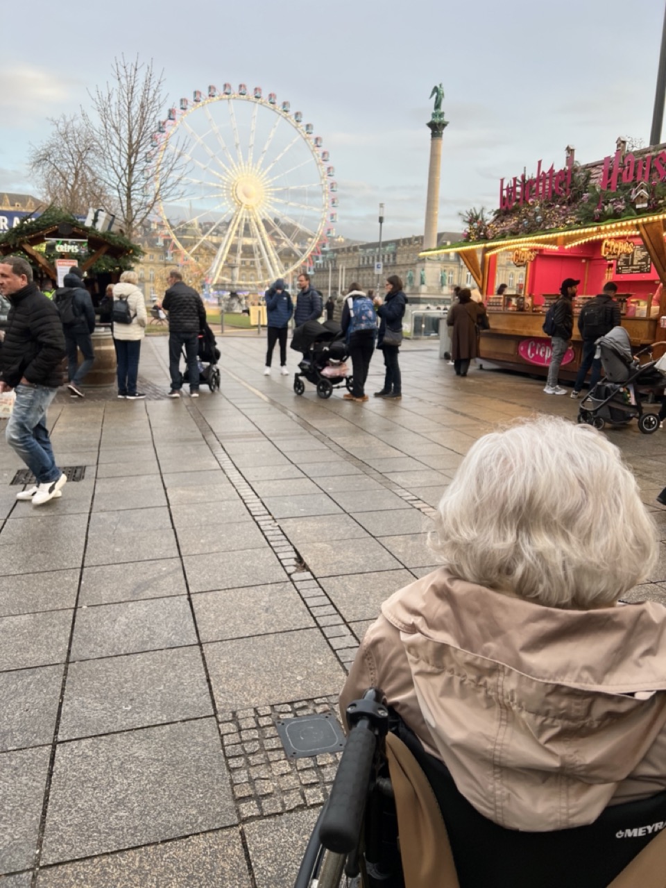 Die von mir begleitete ältere Dame mit weißen Haaren sitzt im Rollstuhl und schaut auf das beleuchtete Riesenrad auf dem Stuttgarter Weihnachtsmarkt.